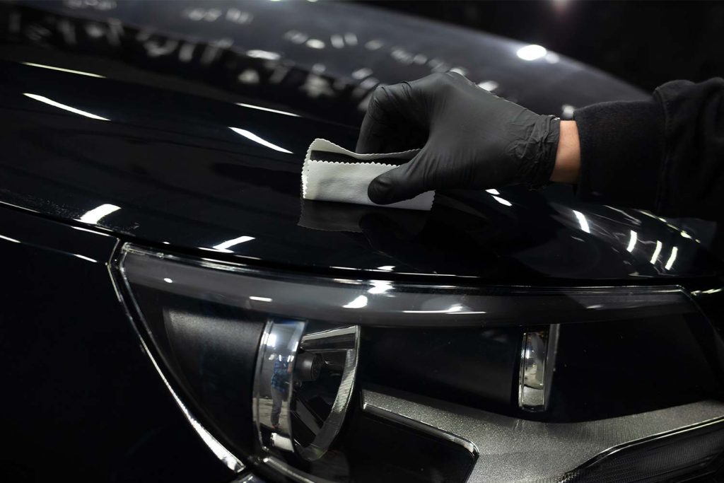 Close-up of a black car’s glossy paint surface being wiped with a microfiber cloth by a person wearing a black glove, highlighting a clean, reflective finish near the headlight.