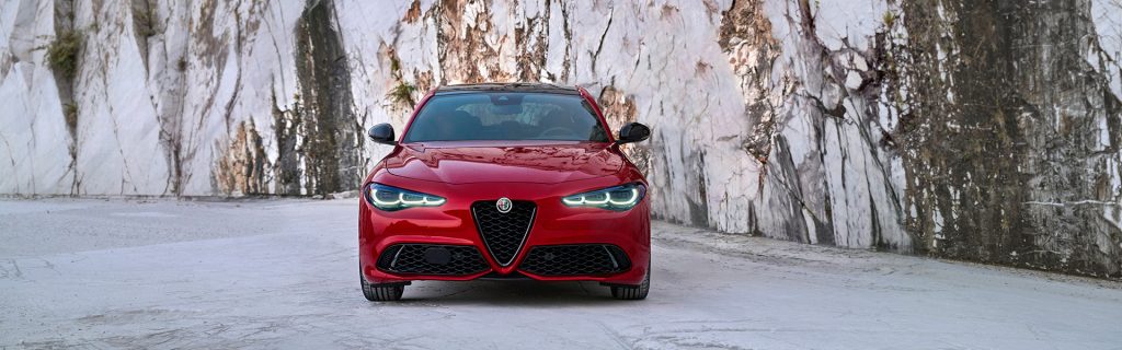 A front view of a red Alfa Romeo Giulia sedan parked on a white stone surface against a dramatic backdrop of a marble quarry cliff wall.