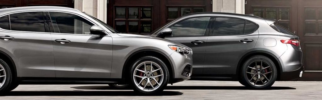 A silver Alfa Romeo Stelvio SUV parked in front of a dark gray Alfa Romeo SUV, showcasing their side profiles and sporty alloy wheels against a background of dark wooden doors.